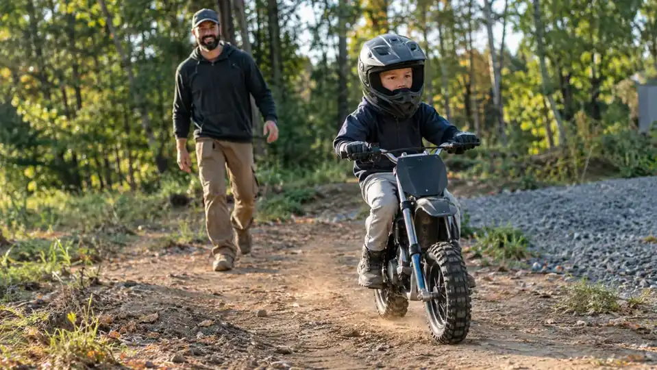 A child in a helmet riding an electric dirt bike along a dirt trail with a parent watching from a few steps behind
