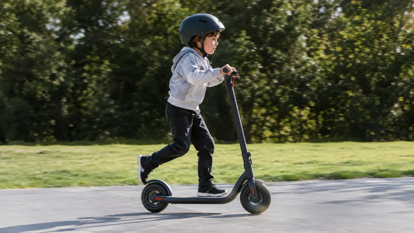 A child in a helmet riding an electric scooter across a smooth concrete pad, grass and a tree line behind them
