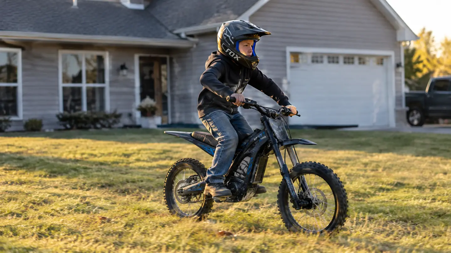 A boy wearing a helmet and long pants rides an electric dirt bike across a flat grass yard beside a house and garage.