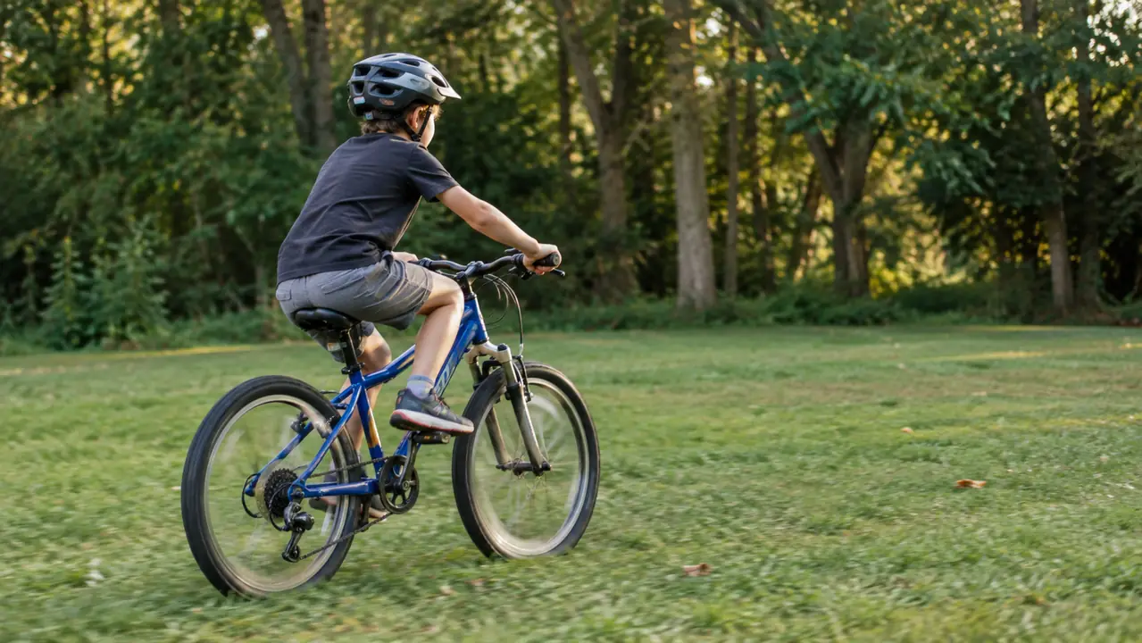 A child wearing a multi-sport helmet riding a bicycle across a flat grass yard near a tree line