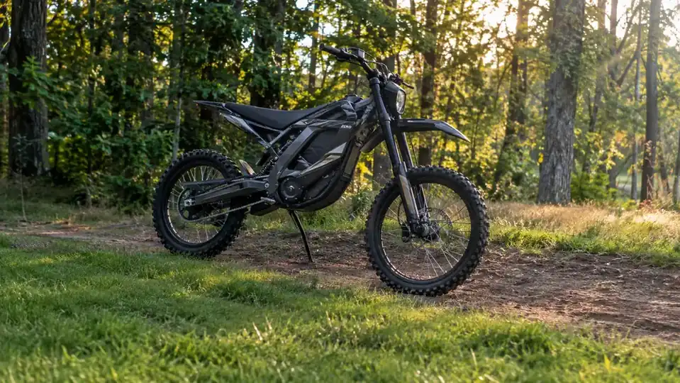 An electric dirt bike standing on a dirt trail at the edge of a yard with trees behind it
